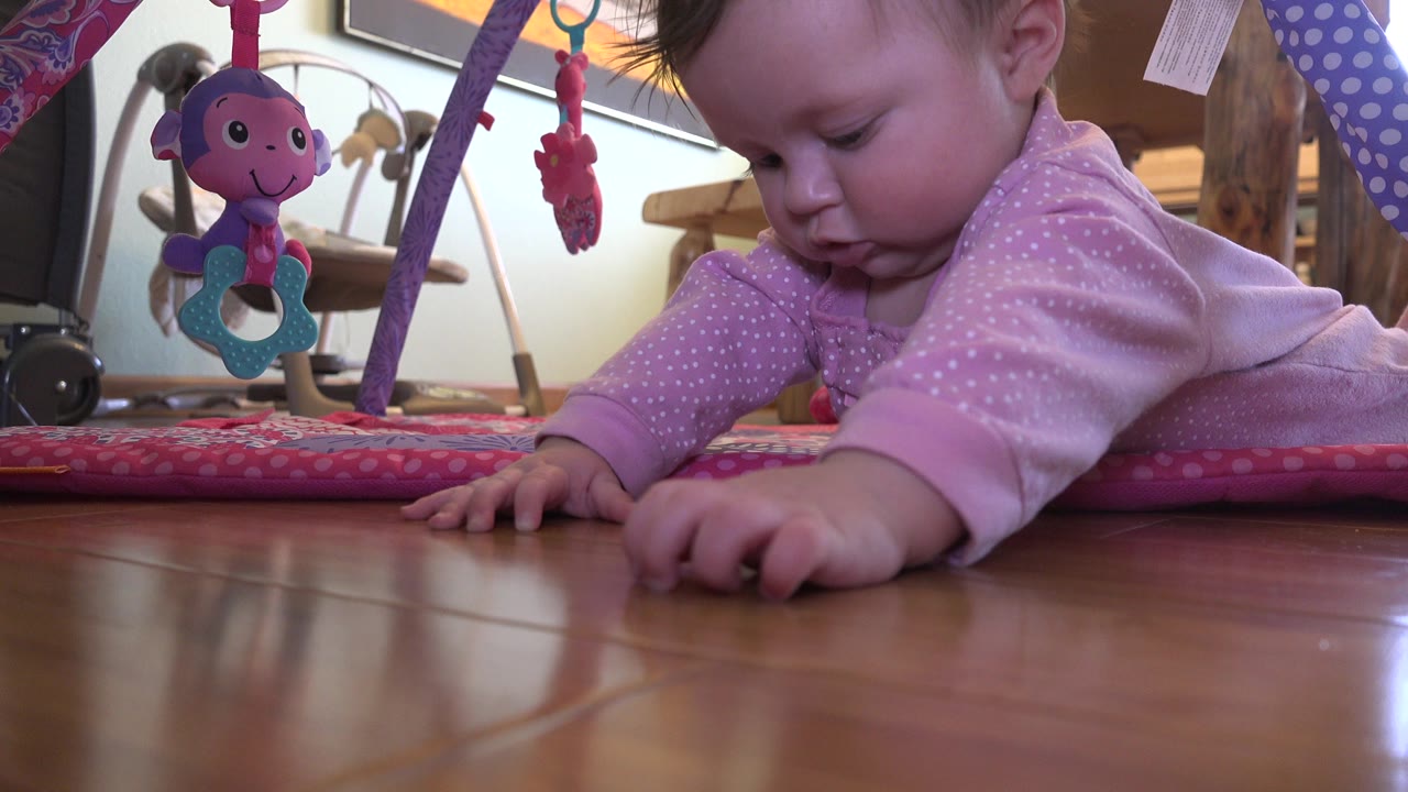 Baby in pink outfit reaching on wooden floor beside hanging toys.