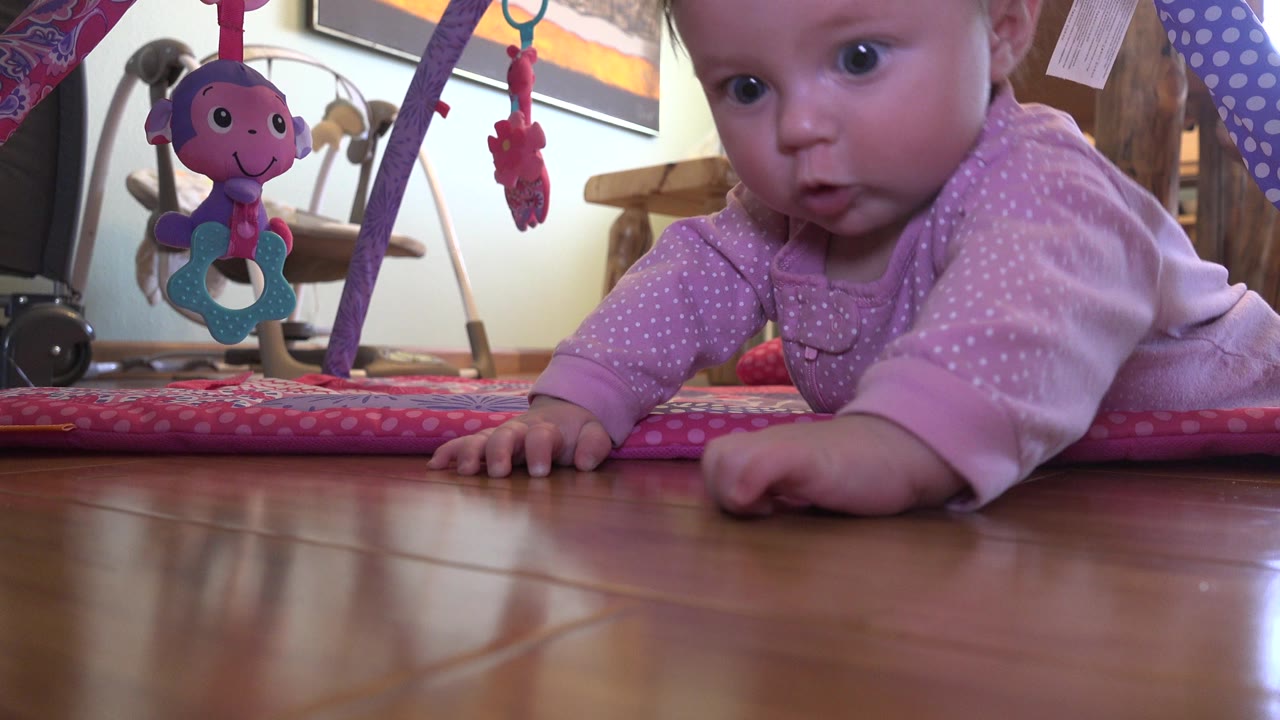 Baby on a wood floor reaching near a pink play mat and hanging toy.