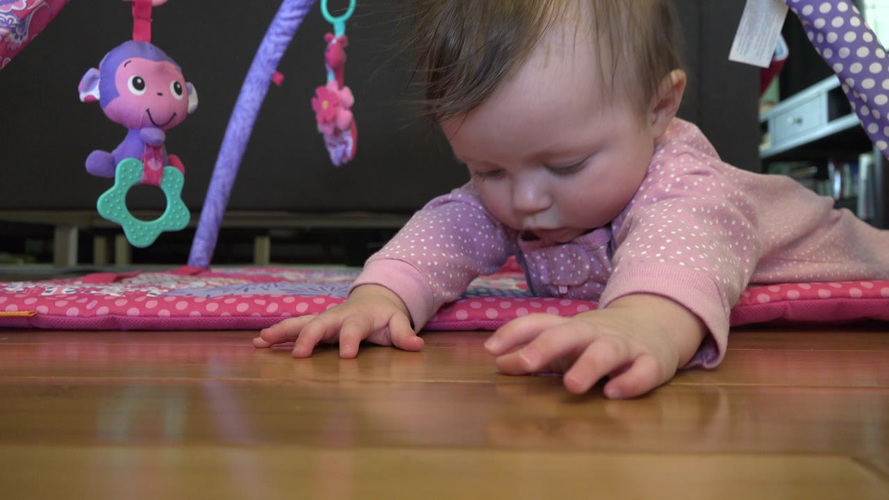 Baby on stomach reaching across a wooden floor under hanging toys.