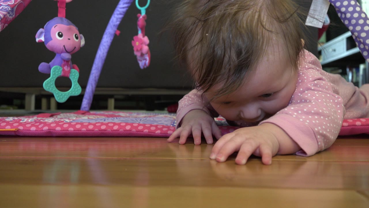 At 0:00, a baby in pink pajamas lies low on a pink play mat in Westminster, Colorado, reaching across a polished wood floor with hanging toys above.