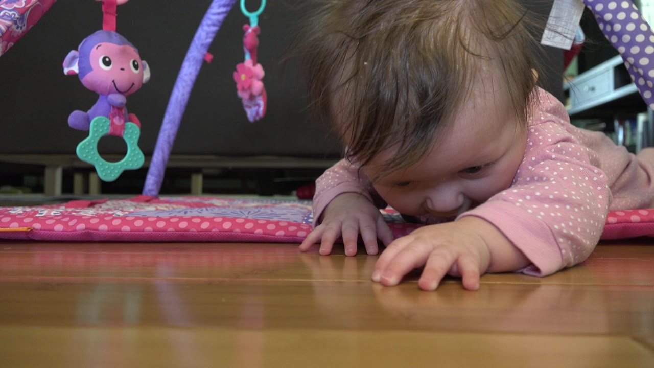 At 0:01, the baby in pink pajamas dips their face closer to the wood floor while lying on the play mat, with a purple plush toy hanging nearby.