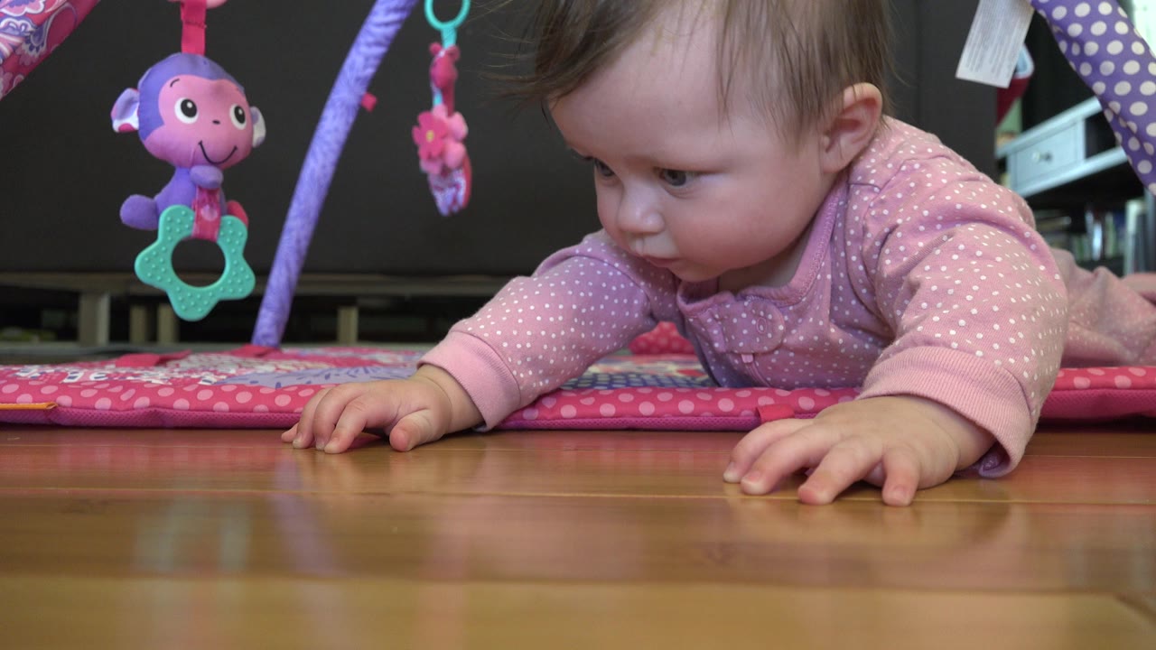 The baby remains prone on the pink play mat in Westminster, Colorado, with one hand extended across the wood floor and a plush toy hanging at left.