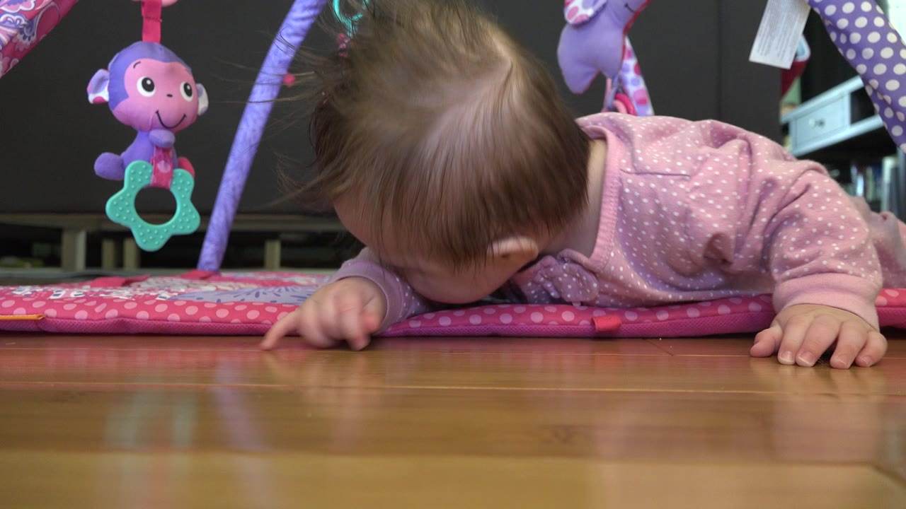 At 0:01 in Westminster, Colorado, the baby remains low on the pink mat, with hair falling forward, a hand on the floor, and a purple toy hanging in the background.