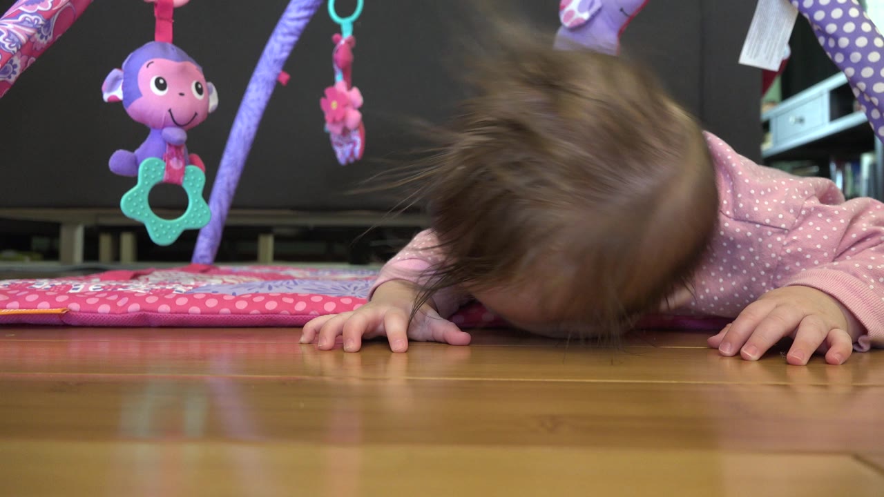 At 0:00 in Westminster, Colorado, a baby lies face-down on a polished wood floor under a pink play mat with purple and teal hanging toys.