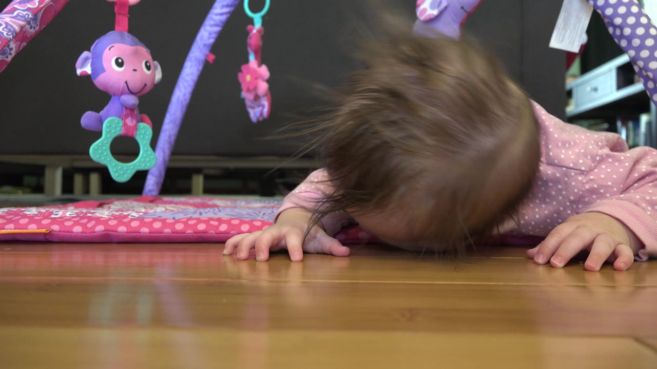 At 0:01 in Westminster, Colorado, the baby remains low on the wood floor with both hands planted while a purple toy hangs above the play mat.