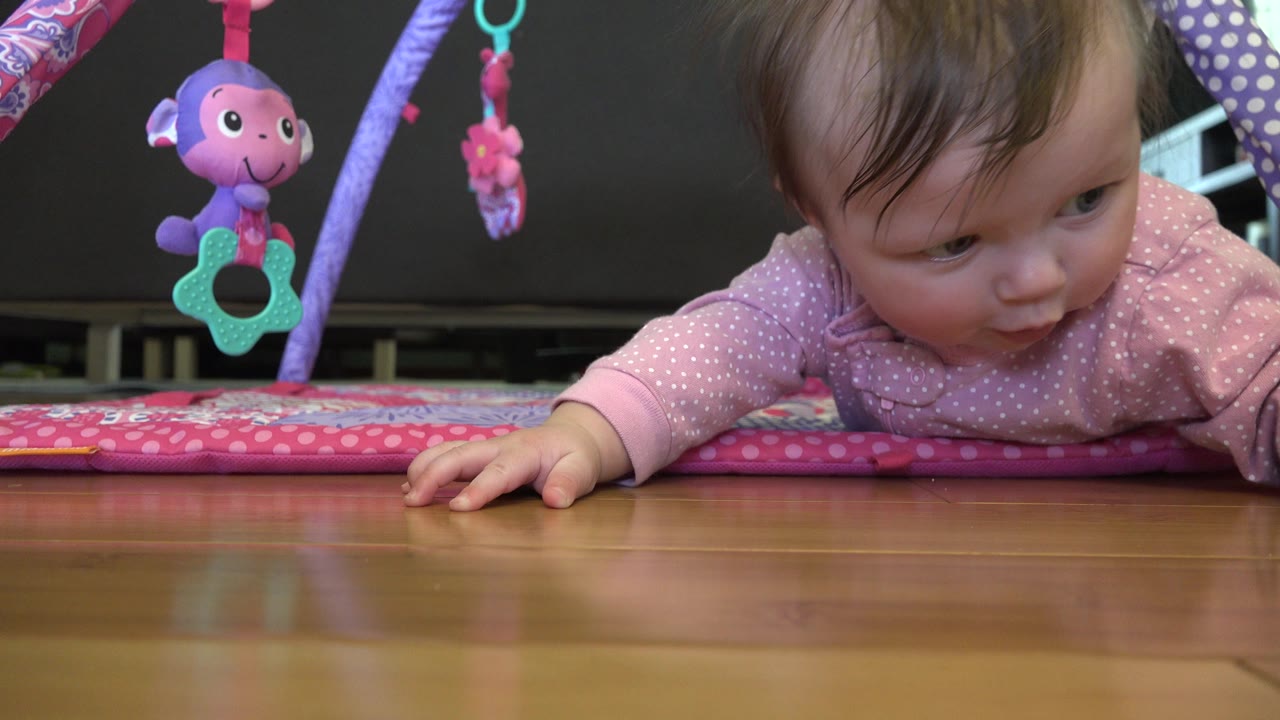 At the start of the Westminster, Colorado clip, a baby lies face down on a pink play mat with one hand on the wood floor and purple hanging toys at left.