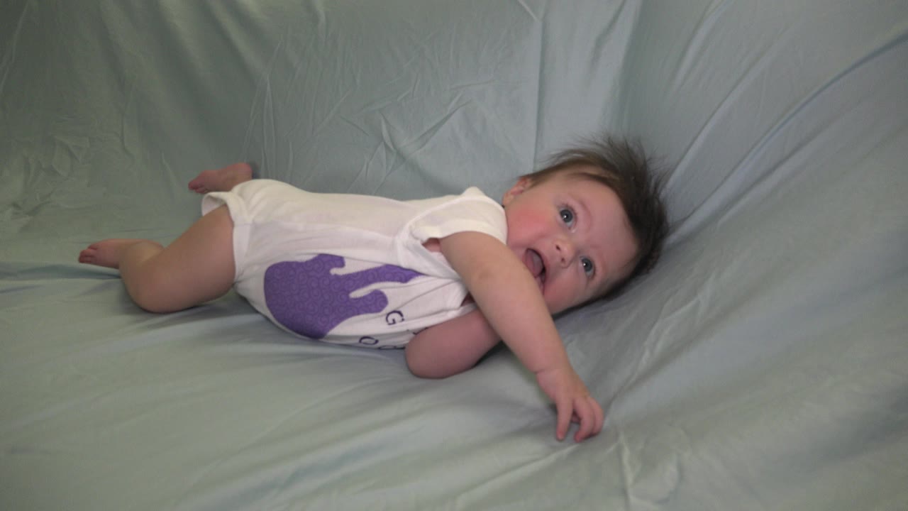 At the start of the Westminster, Colorado clip, a baby girl lies sideways on a pale blue-green sheet with one arm extended and her face turned toward the camera.
