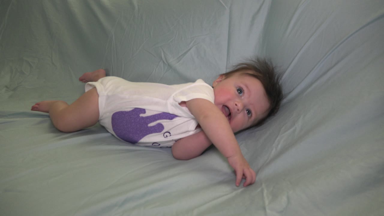 The baby girl remains on her side in Westminster, Colorado, reaching one hand forward across the wrinkled pale sheet while looking up toward the camera.