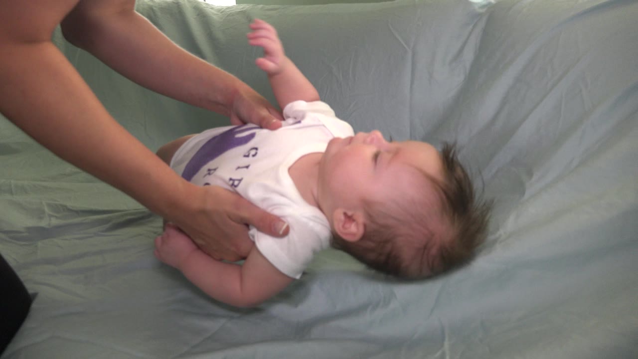 Adult hands support a baby lying on pale bedding.