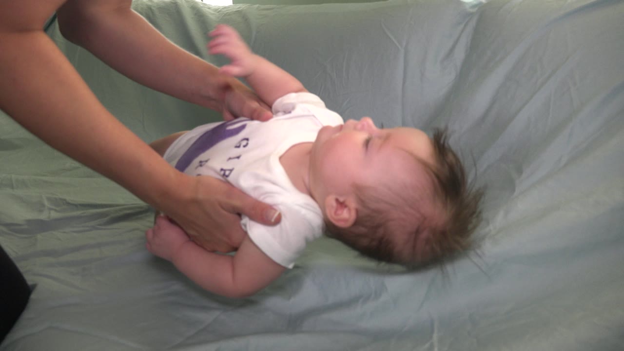 In Westminster, Colorado, the infant remains reclined on the pale covering while adult hands hold the baby's sides and one small arm lifts upward.