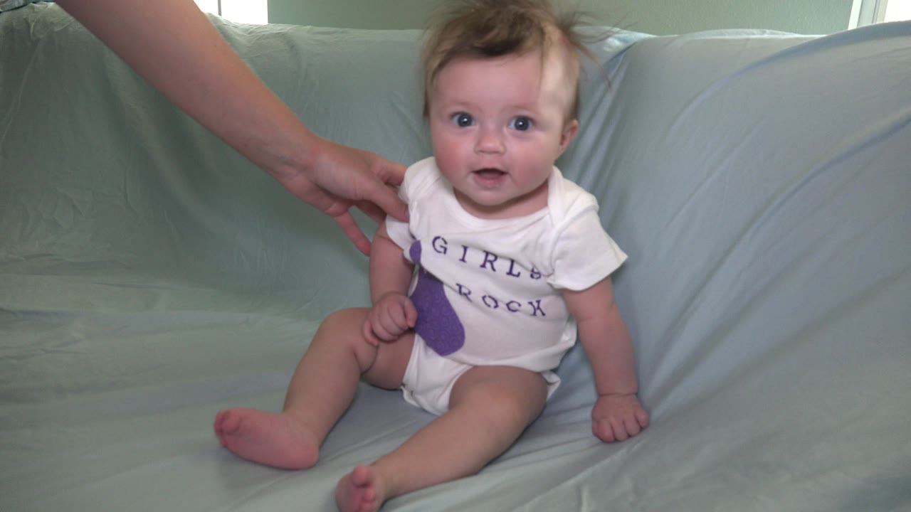 Baby seated on pale blue couch with an adult hand nearby.