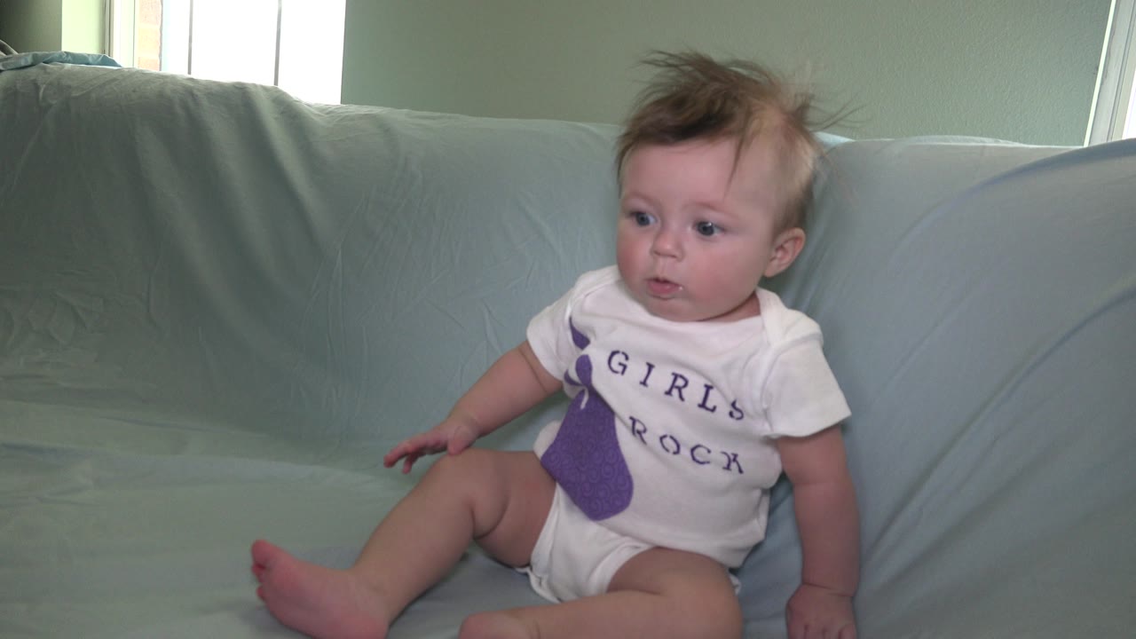 Baby sitting on a pale blue couch beside a bright window.