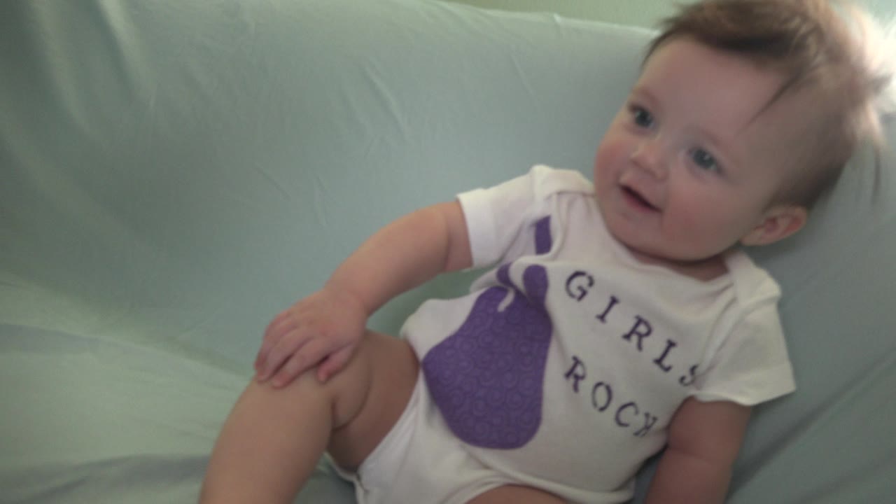 Baby in a white onesie sitting on a pale blue couch.