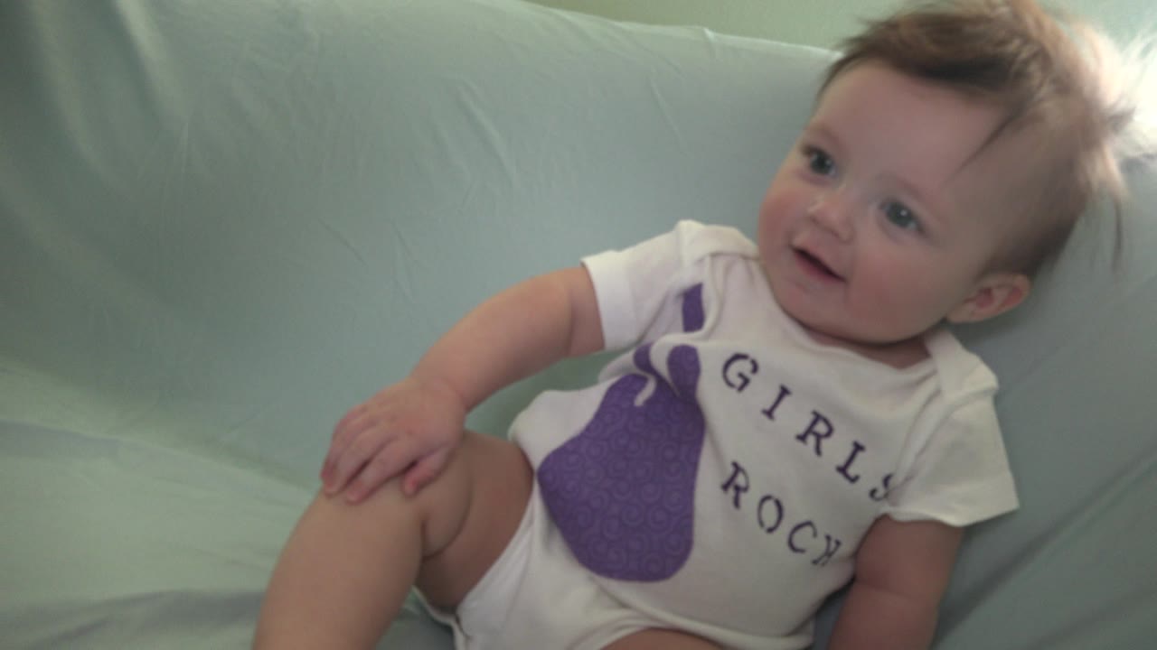 At 0:01 in Westminster, Colorado, the baby leans slightly left on the pale blue couch, one hand near the leg, with the Girls Rock text partly visible on the onesie.