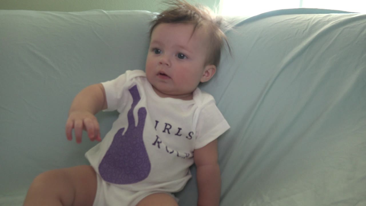 A baby in Westminster, Colorado sits on a pale blue sofa wearing a white onesie with a purple guitar graphic, looking off frame at the start of the clip.