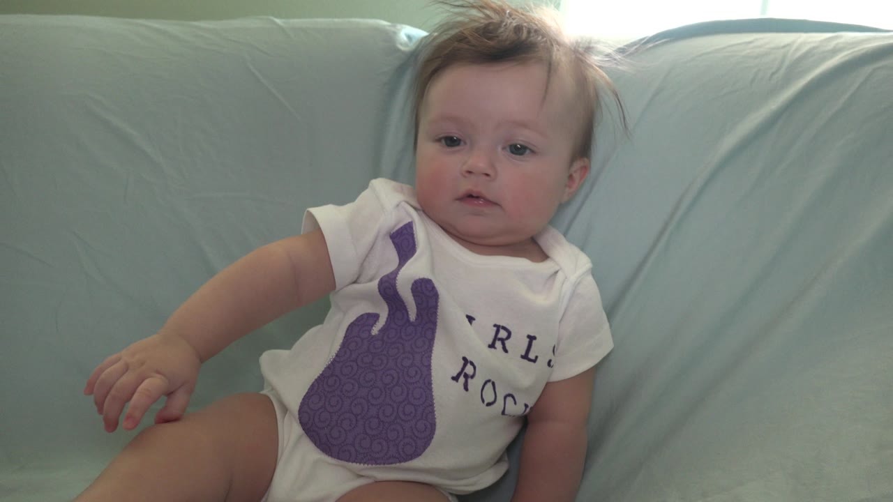 At 0:00 in Westminster, Colorado, a baby with tousled hair sits angled on a pale blue couch, wearing a white shirt with a purple guitar graphic and partial lettering.
