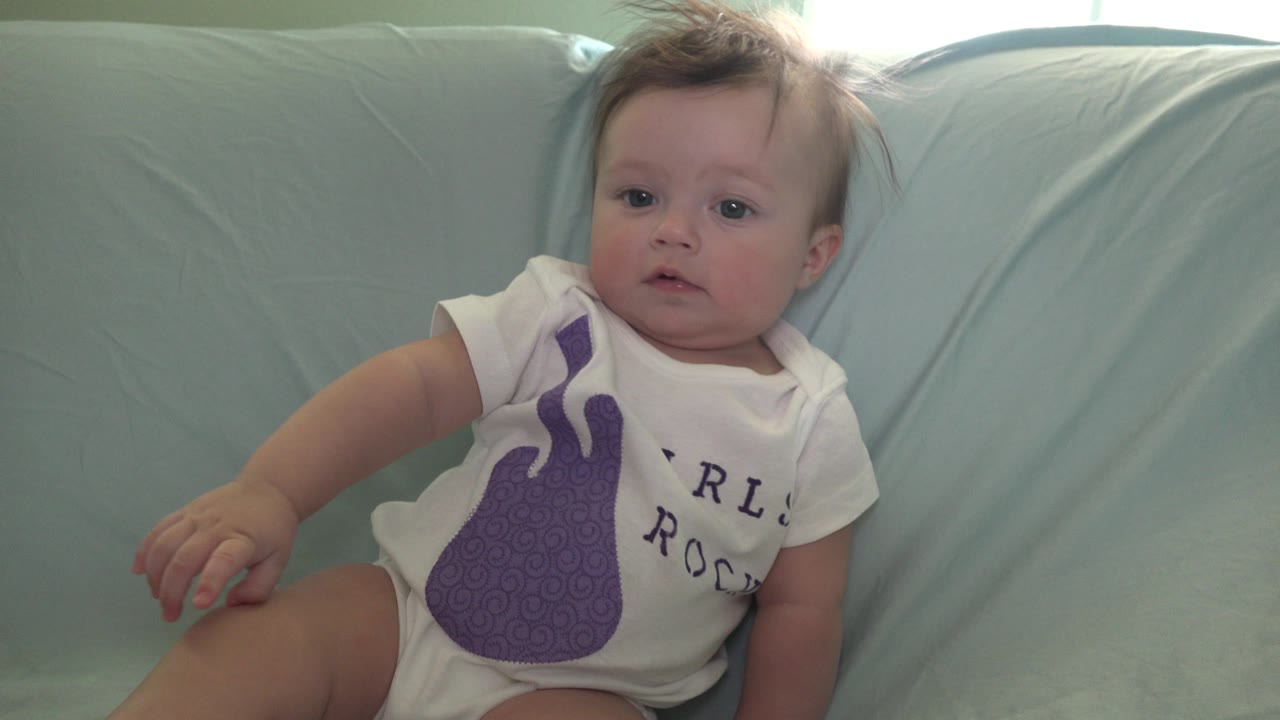At 0:01 in Westminster, Colorado, the baby remains reclined against the pale blue couch, looking forward with one hand near a bent leg and the purple shirt graphic visible.