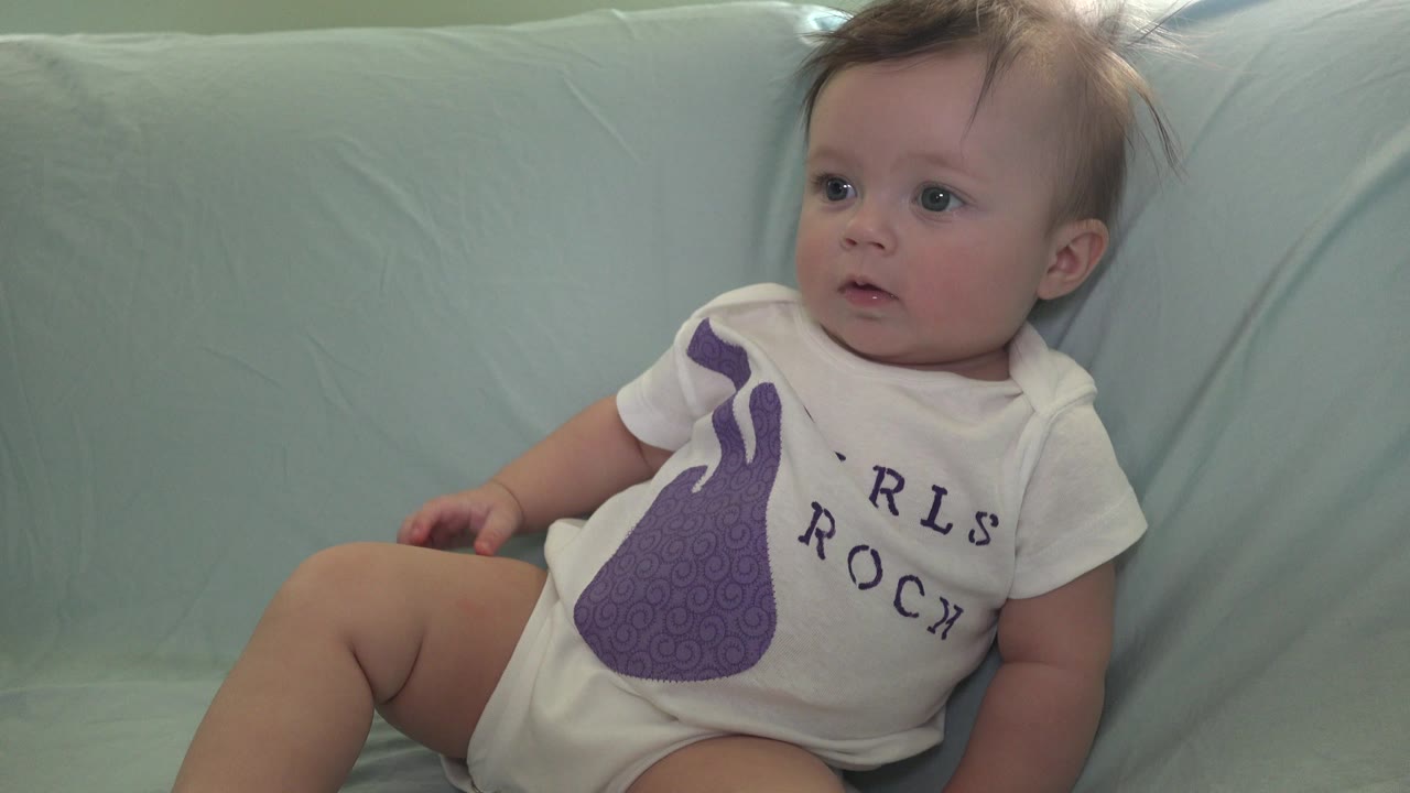 The baby remains seated on the pale aqua sofa in Westminster, Colorado, with a steady side gaze and one hand near a bent knee.