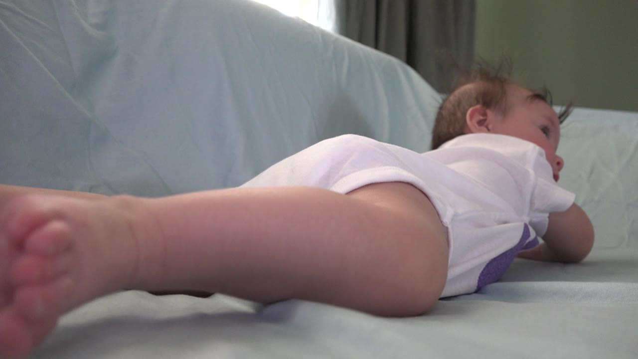 At 0:01, the baby remains on the couch in Westminster, Colorado, with a bare leg reaching across the foreground and the infant's face turned partly to the side.