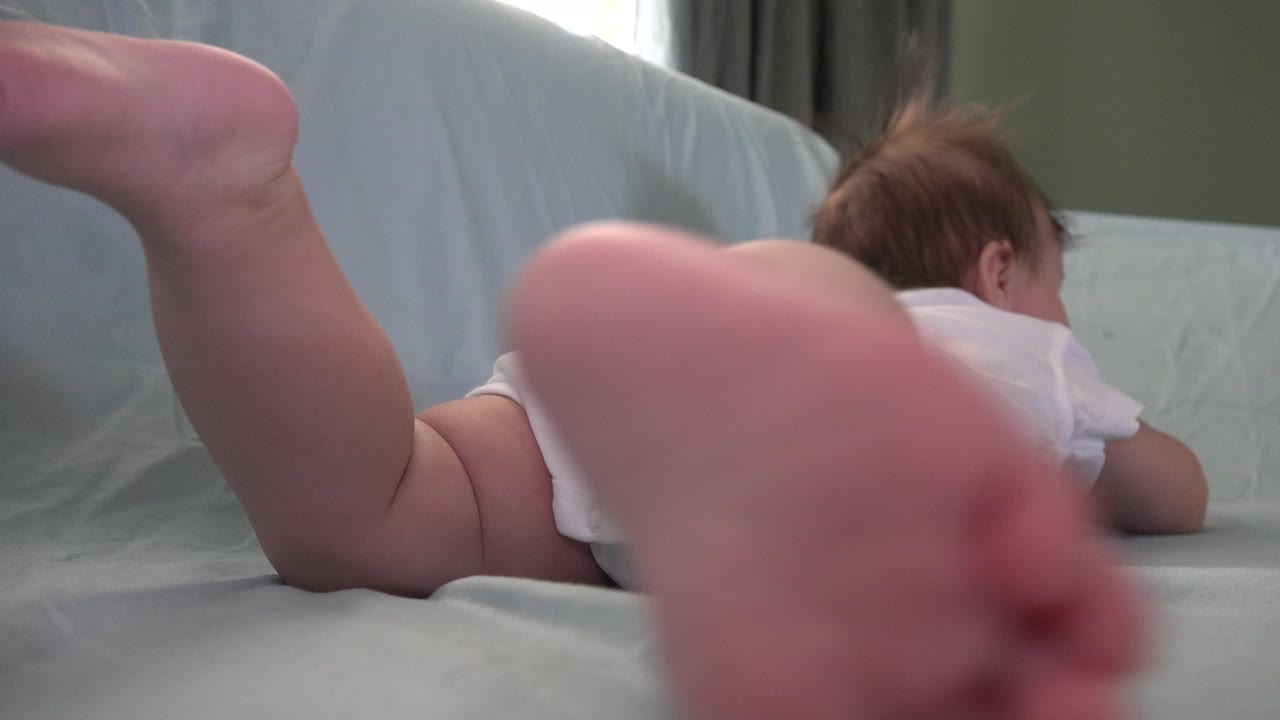 At 0:01 in Westminster, Colorado, one of the baby's bare feet fills the center foreground while the baby remains face down in a white top on the pale sheet.