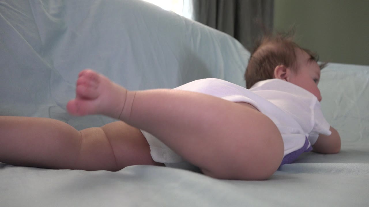 At 0:00 in Westminster, Colorado, a baby in a white outfit lies on a pale blue covered couch or sheeted surface with one bare foot lifted toward the low camera.