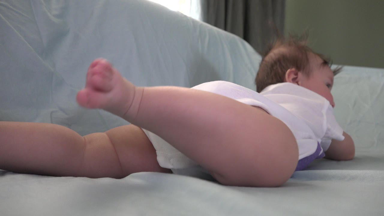 At 0:01 in Westminster, Colorado, the baby's leg stretches across the foreground while the infant faces away on a pale blue covered surface under soft indoor light.