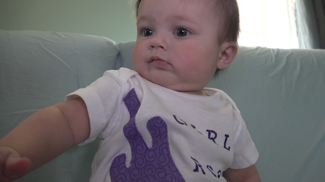 A baby in Westminster, Colorado sits close to the camera on a pale couch, looking to the left with a green wall and bright curtained window behind.