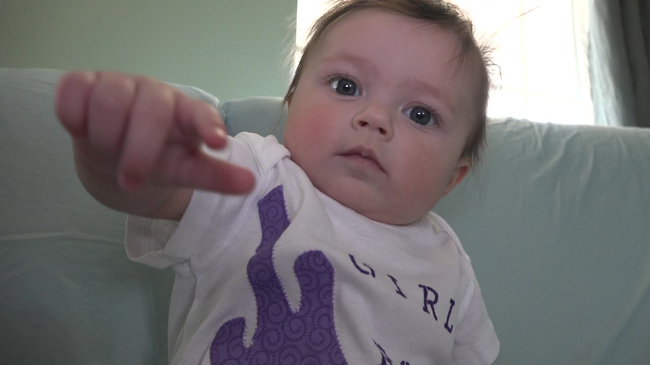 A baby in Westminster, Colorado looks toward the lens while a small hand reaches into the upper foreground, with couch cushions and a bright window behind.