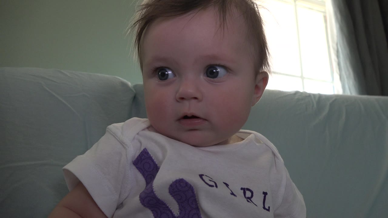 Baby with wide eyes seated on a pale couch near a bright window.