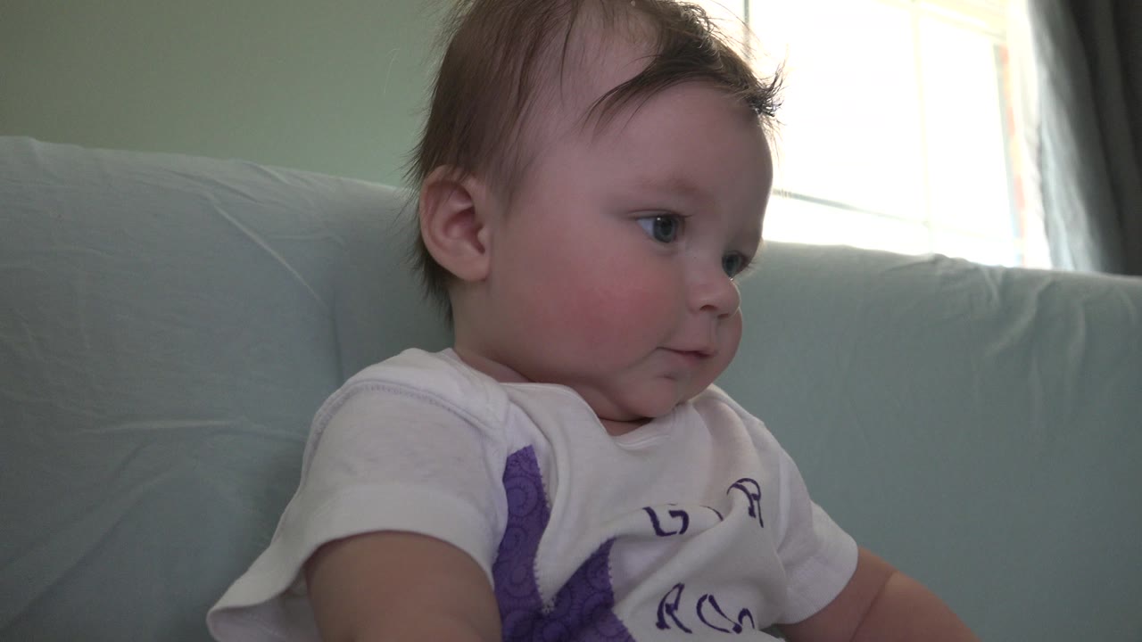 Baby in a white shirt looking left while seated against a pale blue couch.