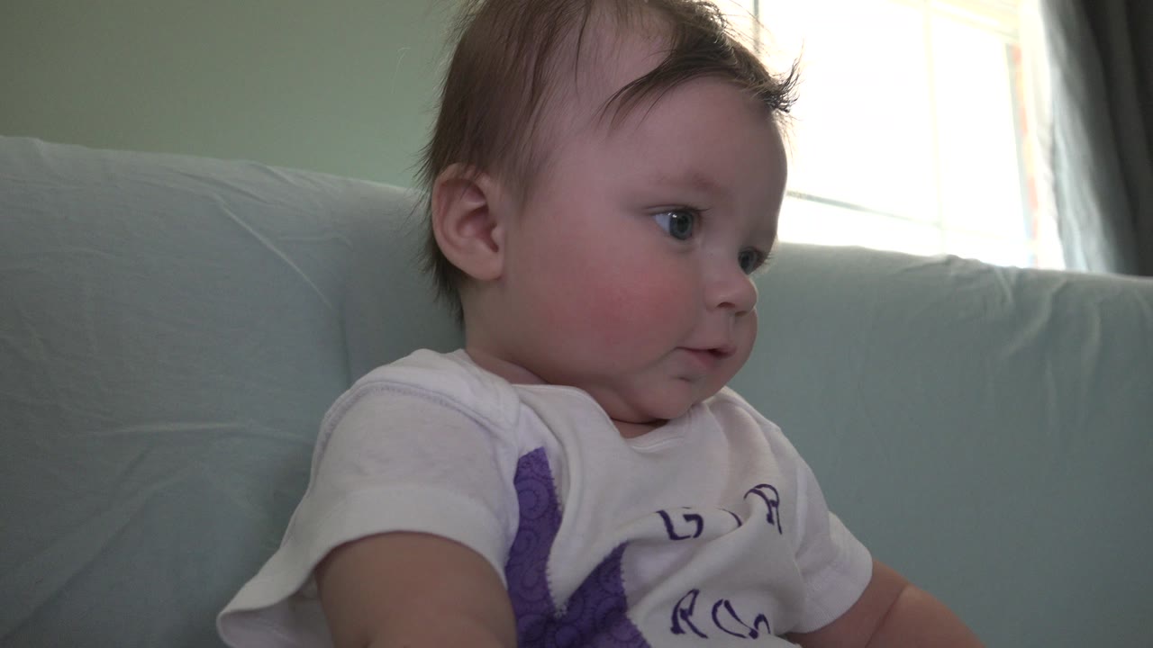 The baby remains turned left in the Westminster interior, framed tightly against the couch with soft backlight from the window.