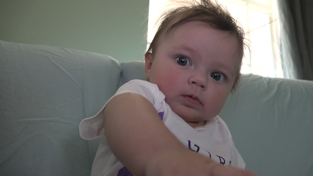 Baby leaning near a pale blue sofa with a bright window behind.