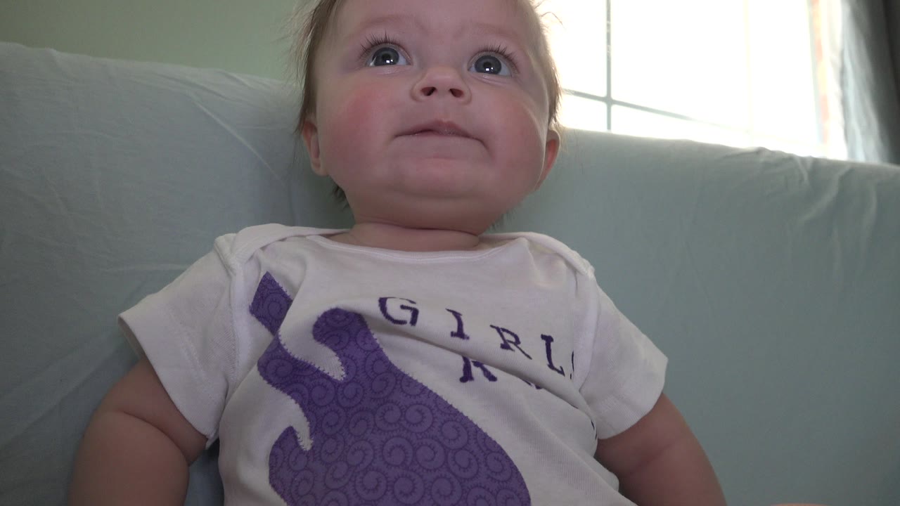 A baby in Westminster, Colorado sits close to the camera on a pale blue sofa, looking upward with a bright window behind.
