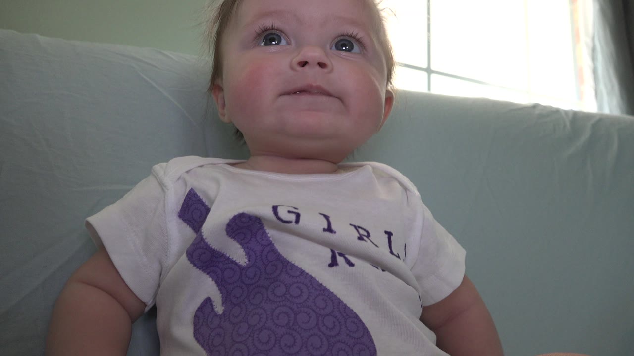 The Westminster, Colorado home clip holds on the baby seated against the sofa, with the face turned upward and daylight from the window at the rear.
