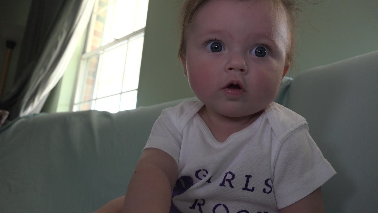 Baby seated close to camera on a pale sofa beside a bright window.