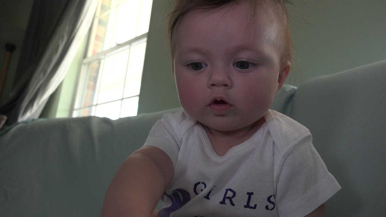 At 0:02, the baby looks down and reaches forward on the pale sofa, with the bright window, curtains, and muted green wall still visible behind in Westminster, Colorado.