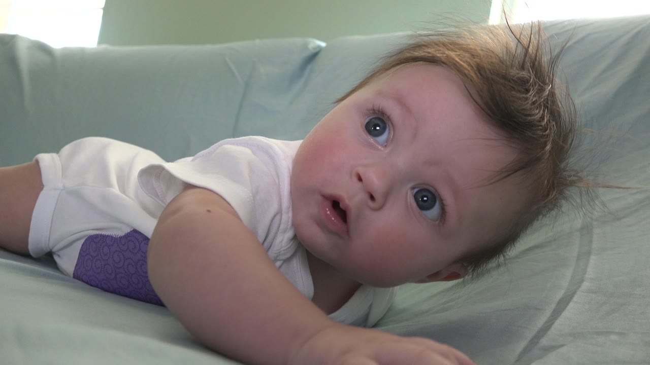 In Westminster, Colorado, the baby remains close to the camera with wide light-colored eyes, a white shirt, and tousled hair against the pale cushion.