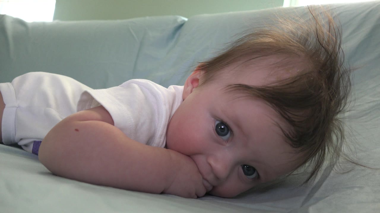 Baby lying on a pale bed with cheek resting on an arm.