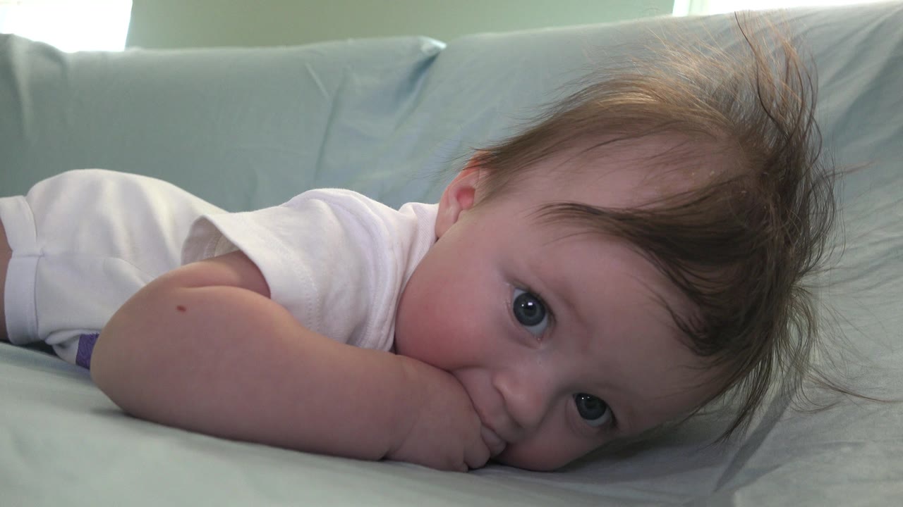 The baby remains prone on the pale bedding in Westminster, Colorado, with wide eyes visible beneath messy hair and pillows filling the background.