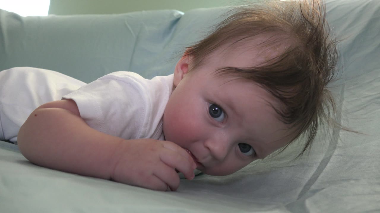 In Westminster, Colorado, the infant lifts one hand toward the mouth while lying on the bed, with both eyes visible and pillows behind.