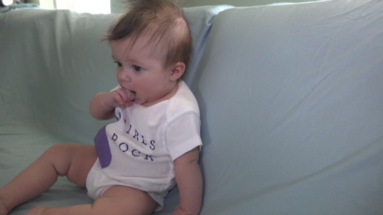 Baby sitting on a pale couch with fingers in mouth.