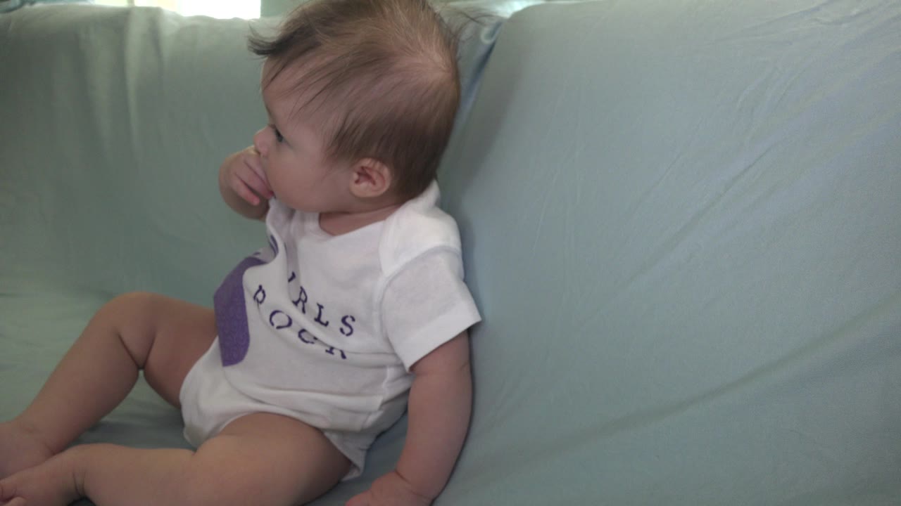 Baby in a white shirt sitting on a pale green sofa with fingers near mouth.