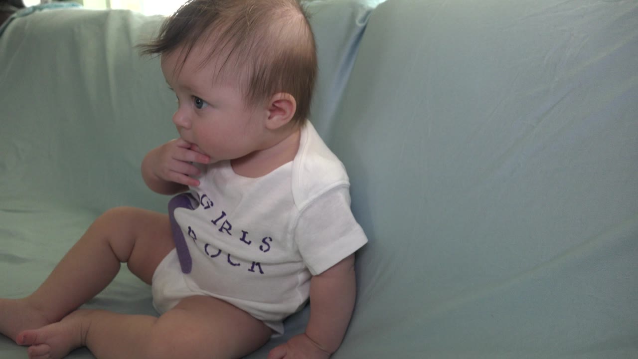 At 0:00, a young child sits on a pale blue-green cushion in a Westminster, Colorado home setting, looking left with fingers near the mouth.