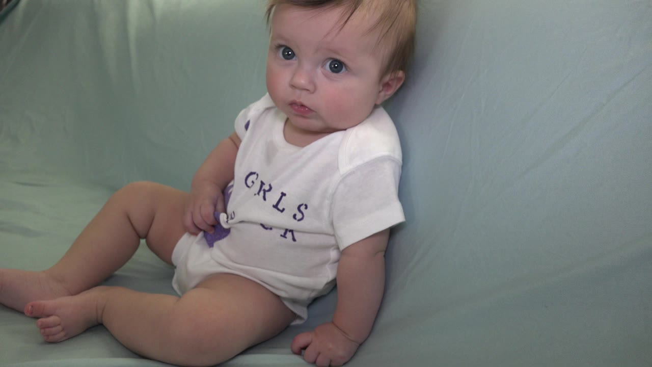 Baby sitting on a pale green couch in a white onesie.