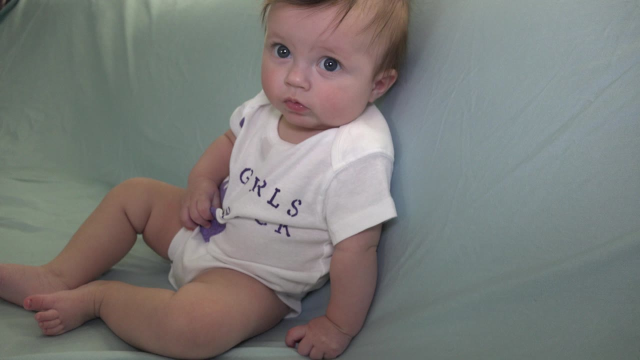 At 0:01, the baby remains seated on the pale green couch in Westminster, Colorado, with one hand near a small purple object and eyes turned toward the camera.