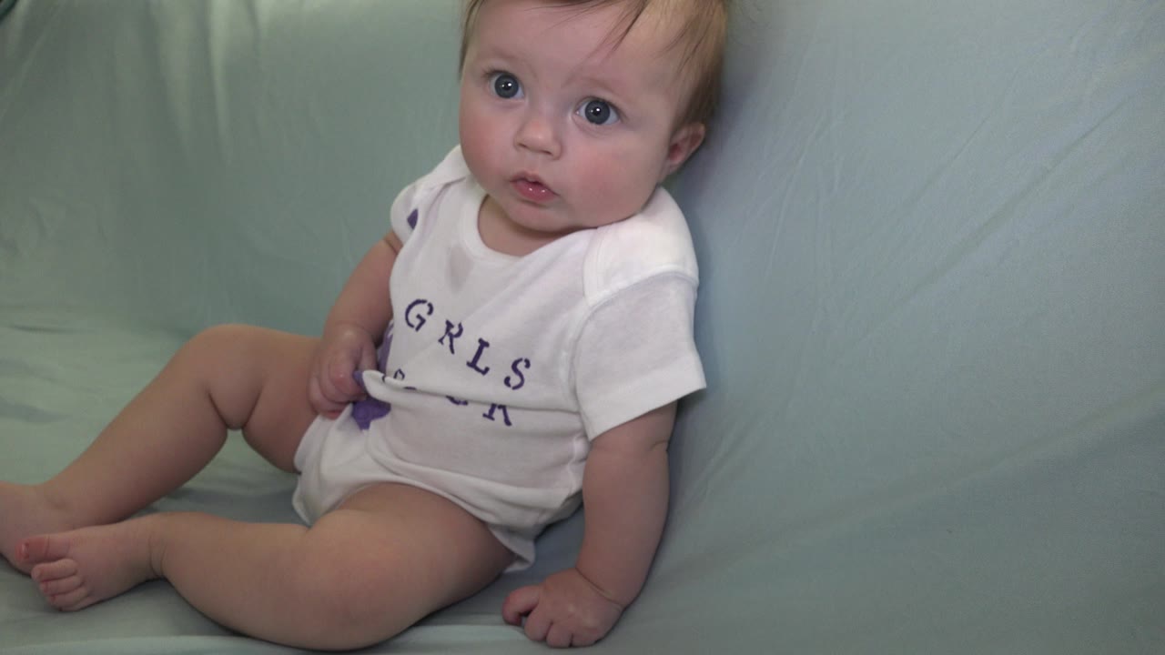 At 0:02, the baby sits upright on the pale green couch in Westminster, Colorado, looking toward the camera with the white onesie lettering partly visible.