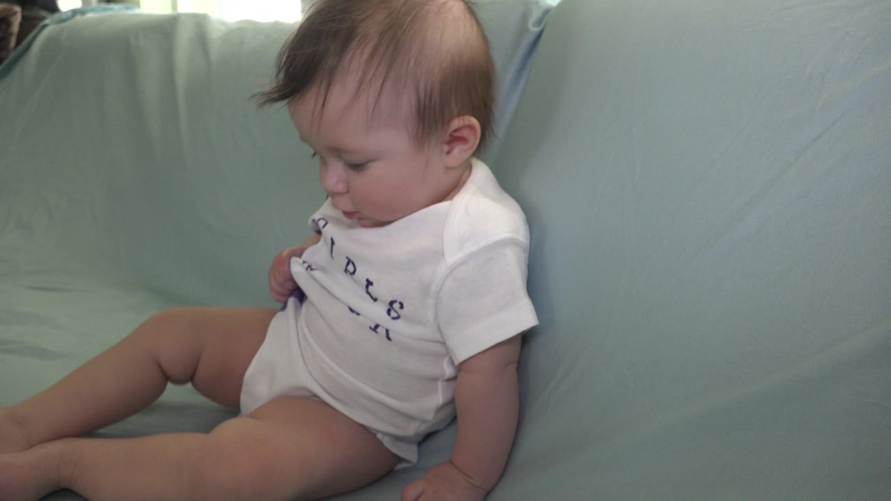 At 0:00, an infant in a white shirt sits on a pale blue sofa in an indoor Westminster, Colorado home setting, looking downward with legs stretched across the cushion.