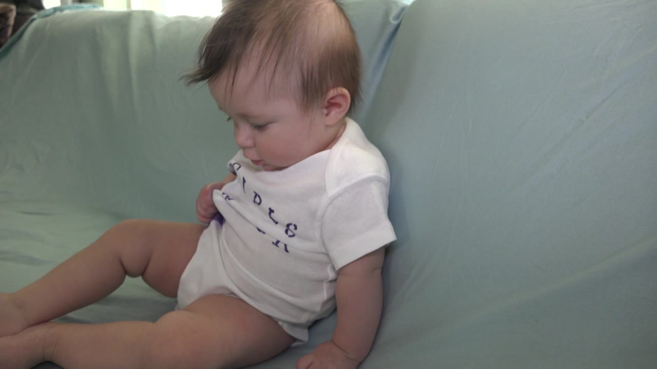 At 0:01, the baby remains seated on the pale blue sofa in Westminster, Colorado, with one hand near the shirt and the face angled down.