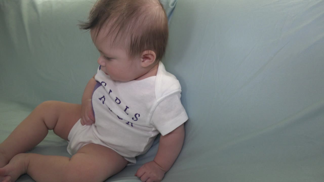 Baby seated sideways on a pale green sofa in a white shirt and diaper.