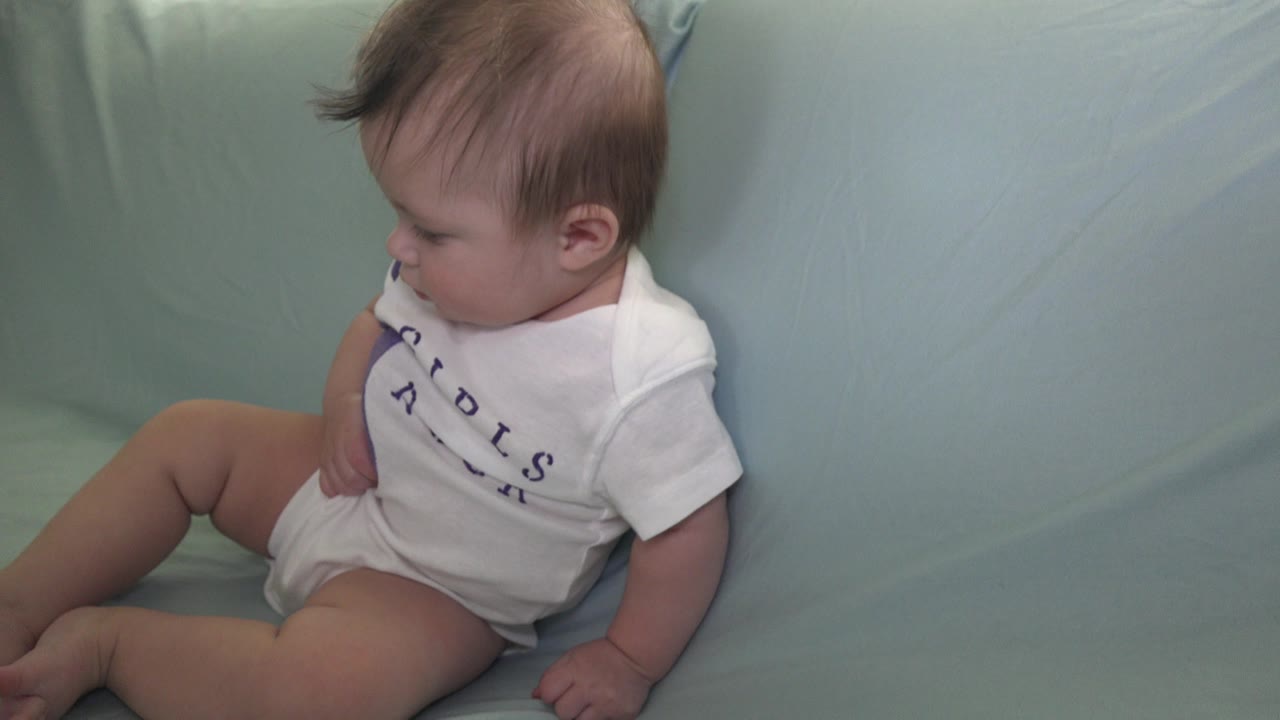 At 0:01, the baby remains seated on the green sofa in Westminster, Colorado, looking down toward a purple cloth or garment near the shirt.